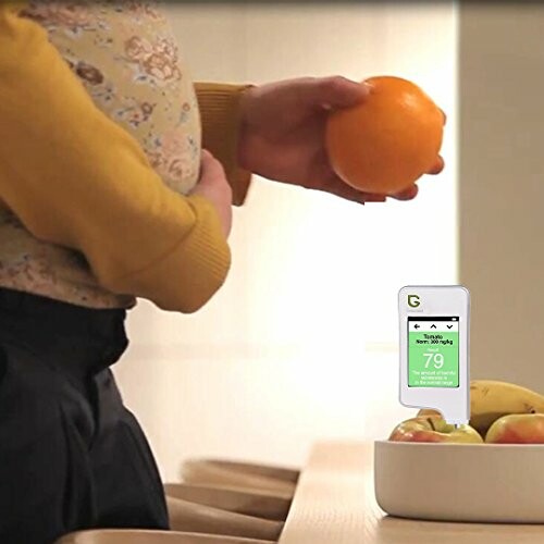 Person holding an orange with a food tester device nearby.